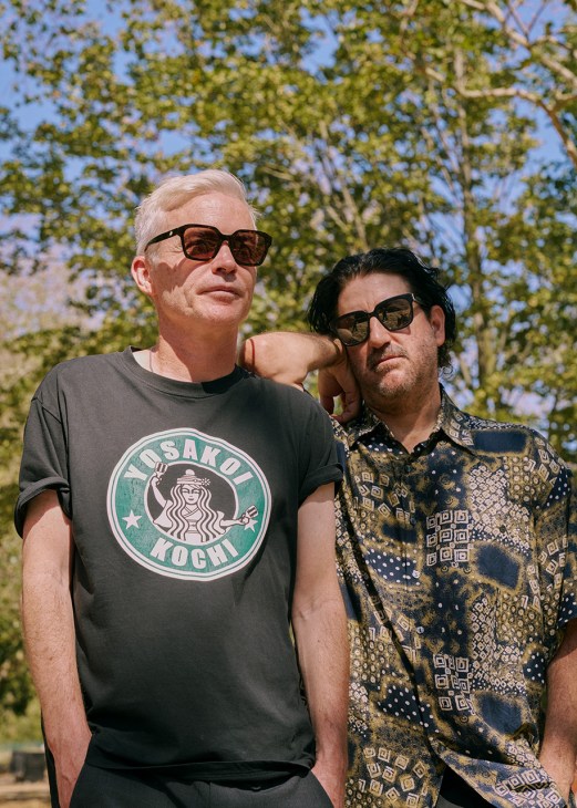 Two men wearing sunglasses stand together outdoors with green foliage behind them.