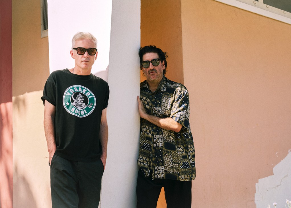 Two men wearing sunglasses stand against a colorful striped wall, one in a black t-shirt and the other in a patterned dark shirt.