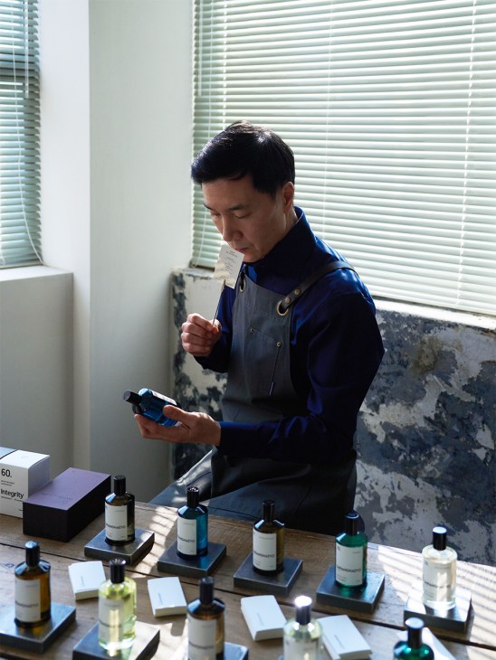 Man in blue jacket examining perfume bottles at a wooden table with multiple Saranghaeyo fragrance samples.