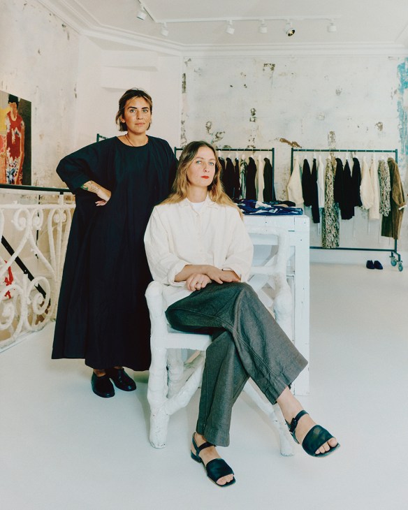 Two women pose in a minimalist shop with distressed white walls and clothing racks displaying garments behind them.