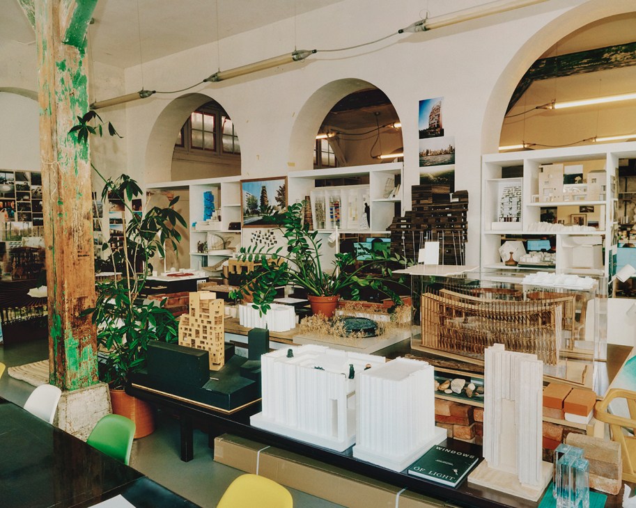 Lina Ghotmeh's studio displaying architectural models, sketches, and reference materials on shelves with plants and exposed column.
