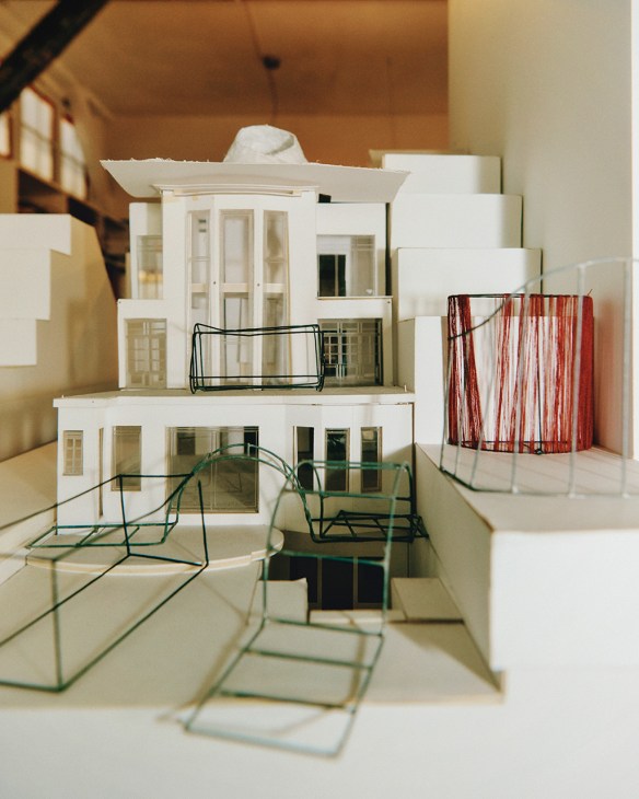 Architectural maquette showing a white multi-story building model with wire furniture details and red fabric accents.