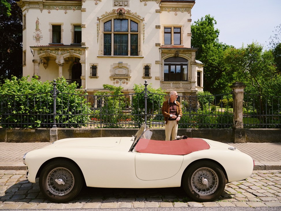Man standing beside a cream-colored 1957 AC Ace Bristol convertible with pink interior in front of an ornate European mansion.