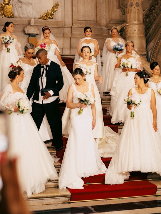 Brides in white wedding dresses and veils posing on grand stone staircase in ornate historic building.