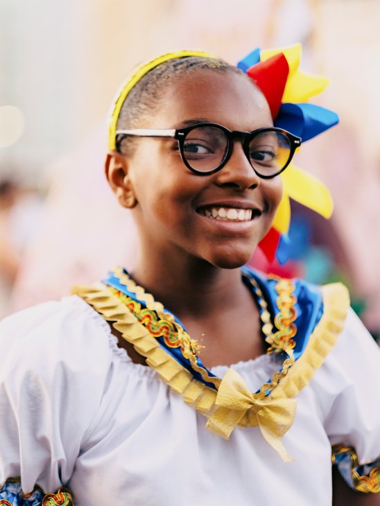 Young child in colorful festival costume with glasses, yellow headband, and decorative collar smiling at Lisbon's marchas populares parade.