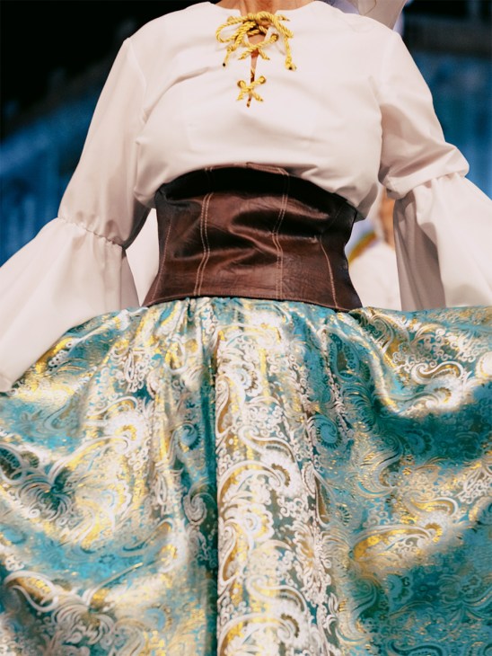 Person wearing a white puffy-sleeved blouse with gold leaf decoration, brown corset, and ornate blue patterned skirt with gold embroidery.