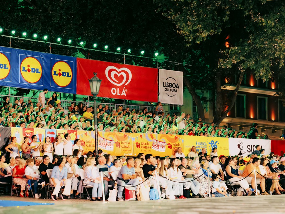 Crowded stands of spectators watching Lisbon's Marchas Populares street festival with sponsor banners overhead.