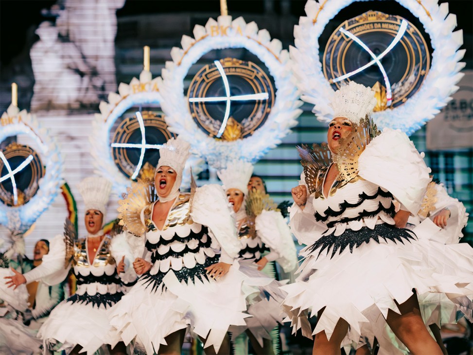 Parade performers in elaborate white feathered costumes with gold accents and black wing-like details, wearing tall white headdresses, dancing in front of large decorative circular backdrops.