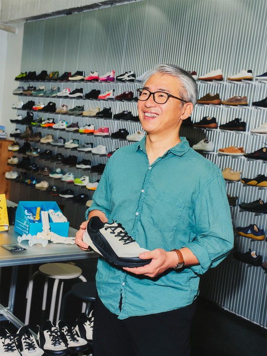 Takashi Fukuchi holds a Notace zero-drop shoe in his Stride Lab flagship store with shelves of footwear behind him.
