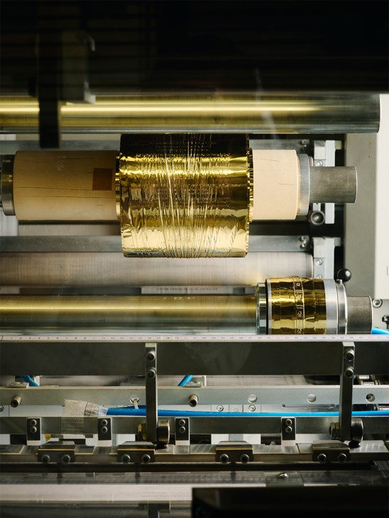 Gold foil roll feeding into a Sonderegger printing machine press with metal rollers and components.