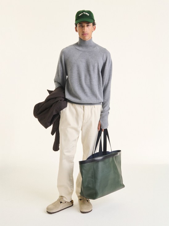 Man wearing gray turtleneck sweater, cream trousers, green cap, and holding a green tote bag