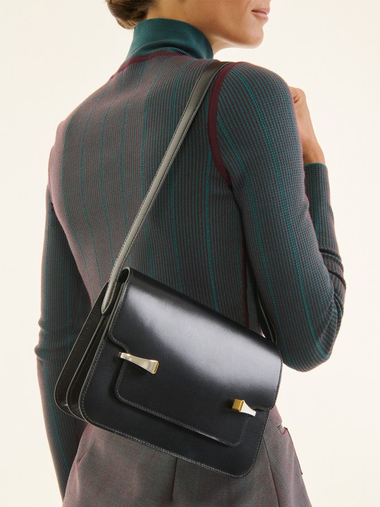 Woman wearing a multicolored knit sweater and carrying a black leather Hermès Clou de Forge shoulder bag with gold hardware.