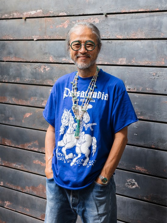 Man wearing blue Paraphobia graphic t-shirt with white elephant design and green beaded necklaces against weathered wooden wall.