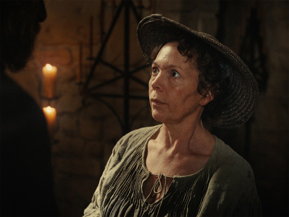 Olivia Colman as an unmarried fisherwoman in medieval fantasy comedy film Wicker, wearing period costume and a netted headdress by candlelight.
