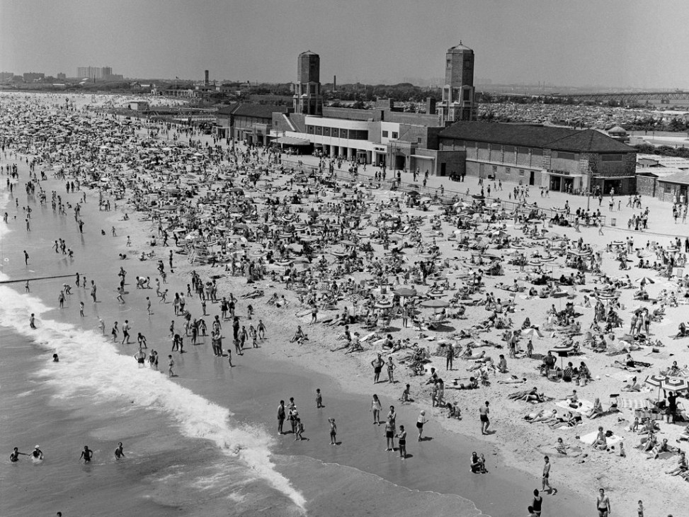Crowded beach scene from the 1930s with bathers and the Jacob Riis Bathhouse art deco building in background.