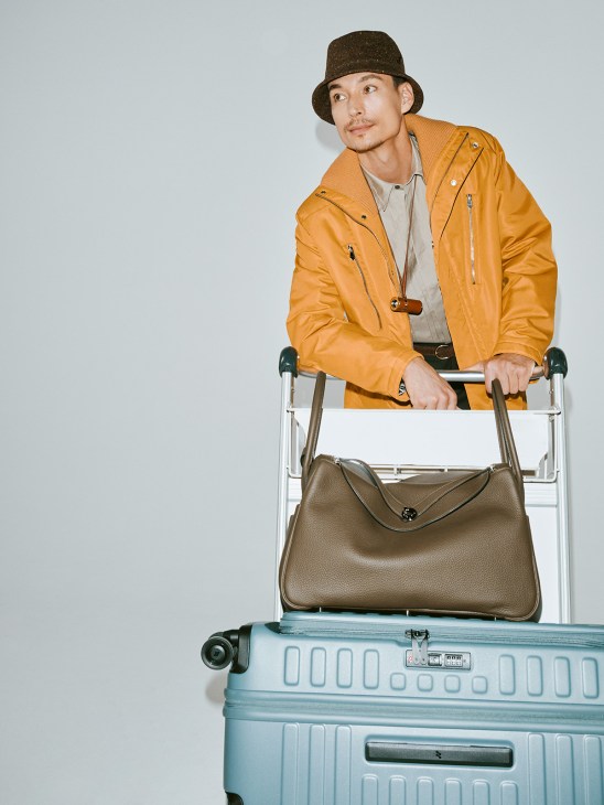 Man in mustard yellow Hermès jacket and brown hat posing with light blue suitcase and brown leather bag.