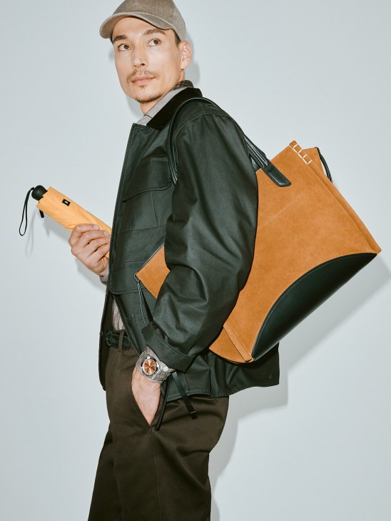 Man wearing Rubato jacket and trousers holding JW Anderson tan leather bag and Blunt umbrella.