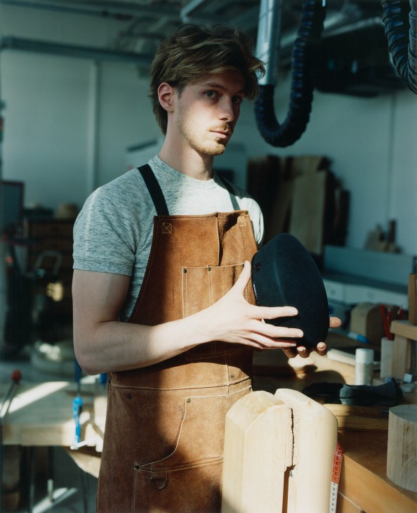 Hatmaker in brown leather apron holding a black felt hat in a workshop