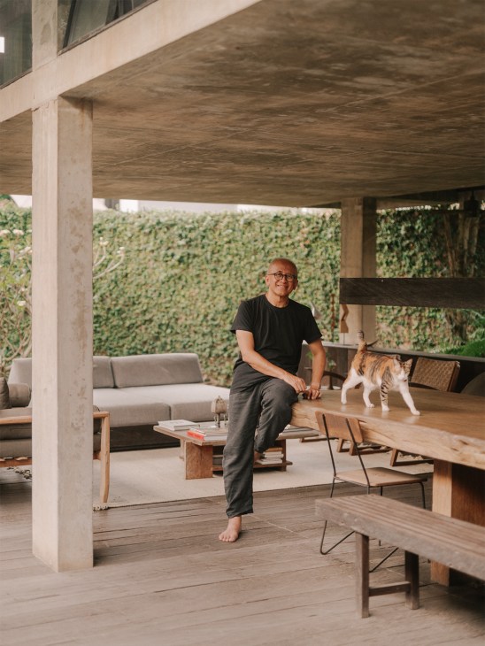 Man sitting on wooden table under concrete overhang in modern residence with pool and green garden visible behind