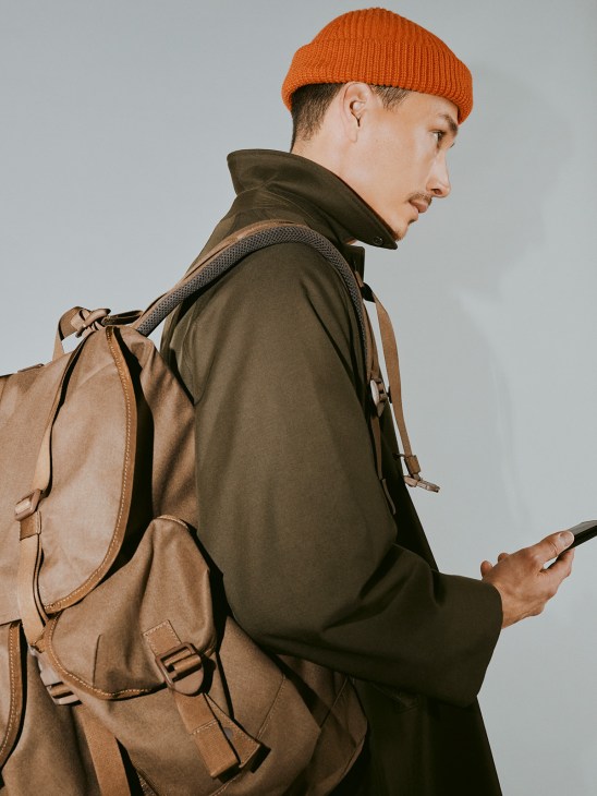 Man wearing olive jacket, orange beanie, and tan leather backpack holding a phone.