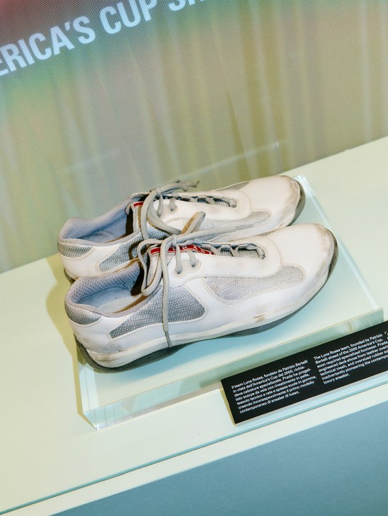 Pair of white and gray Prada Linea Rossa sneakers displayed on a museum-style acrylic platform with an informational plaque.