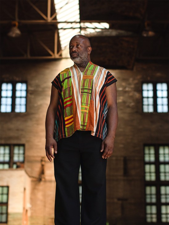 Man wearing colorful striped African textile vest standing in industrial studio with large windows