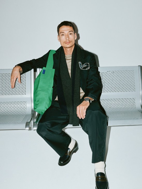 Man in black Ssstein jacket and trousers with green Kaan bag sits on white perforated bench.