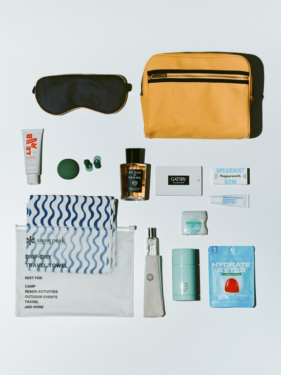 Travel essentials including mustard toiletry bag, eye mask, sunscreen, fragrance, gum, towel, and skincare products arranged on white background.
