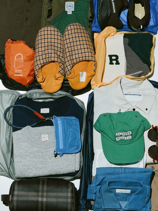Flat lay of travel clothing and accessories including packing cubes, slippers, caps, shirts, bags, and sunglasses.