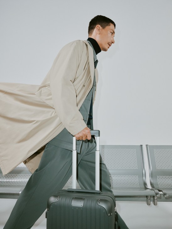 Man in beige Laminar coat and Graphpaper jacket holding Rimowa suitcase at airport gate.