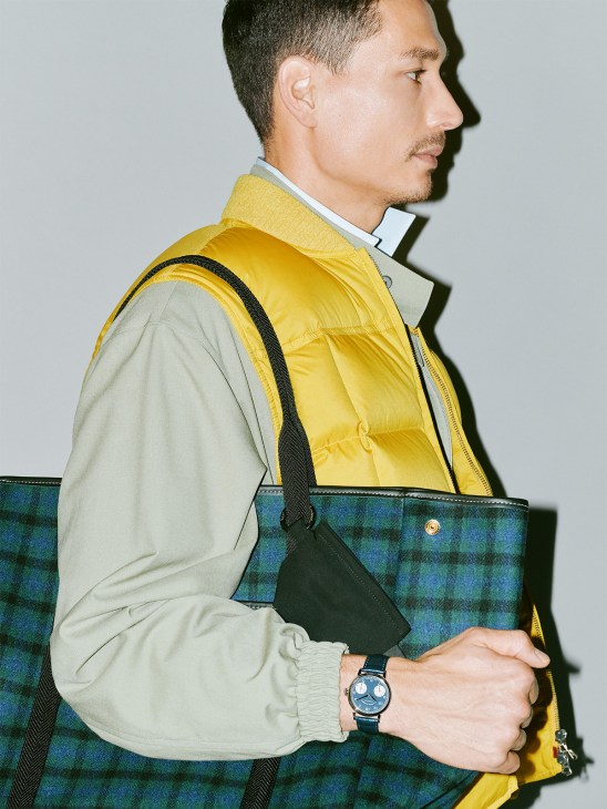 Man wearing yellow puffer vest over light shirt, holding plaid tote bag and small black pouch, photographed in profile