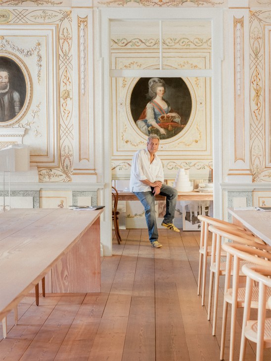 Manuel Aires Mateus sits in his Lisbon architecture office beneath a portrait of Portuguese Queen Maria I, with ornate 18th-century ceiling frescoes and decorative wall paneling visible.