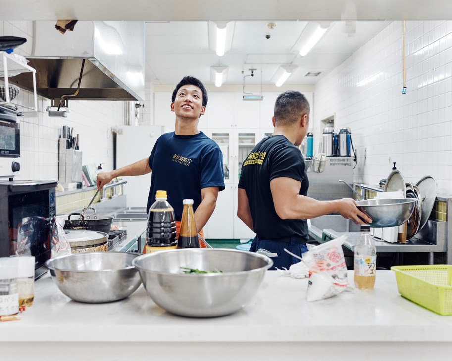 Two Tokyo firefighters in dark uniforms cooking together in a bright commercial kitchen with stainless steel bowls and ingredients on the counter.