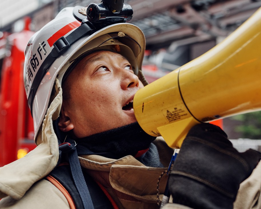 Tokyo firefighter wearing helmet with headlamp uses yellow megaphone to shout commands during training drill.