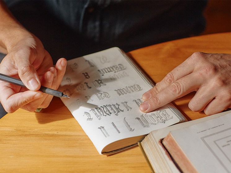 A type designer's hands sketch letterforms in a notebook with pen at a wooden desk.