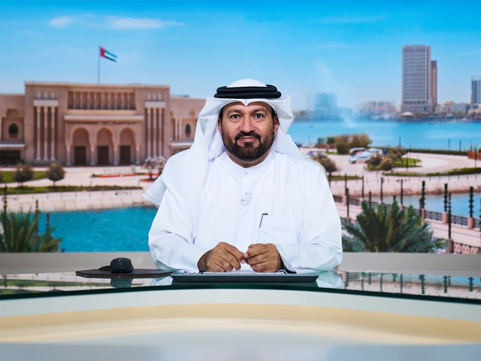 Man in white kandura and ghutra seated at broadcast desk with Sharjah waterfront and pink building behind him.