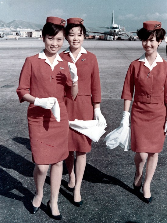 Three female flight attendants in red uniforms with white collars and maroon caps stand on an airport tarmac with a passenger aircraft in the background.