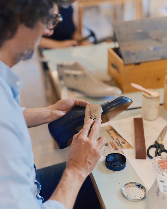 A person wearing glasses and a light blue shirt holds a dark blue shoe form while working at a wooden table with craft materials and containers at Schola Academy.