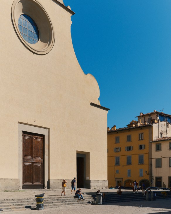 Santo Spirito church facade with circular window and wooden door, piazza with tourists below, colorful buildings adjacent.