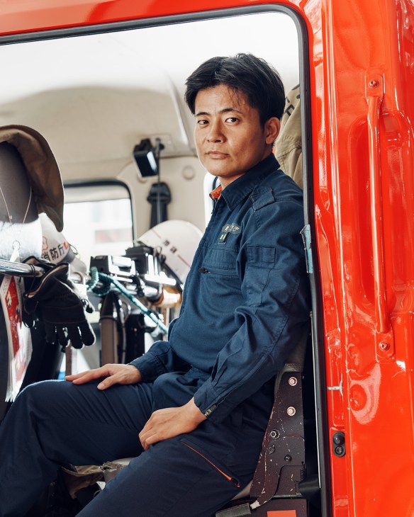 Junior firefighter Genta Saito sits in the cab of a red fire truck wearing a dark blue uniform.