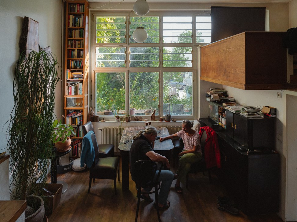 Two people sitting at a table in a modernist home interior with large windows overlooking greenery, bookshelves, and pendant lights.