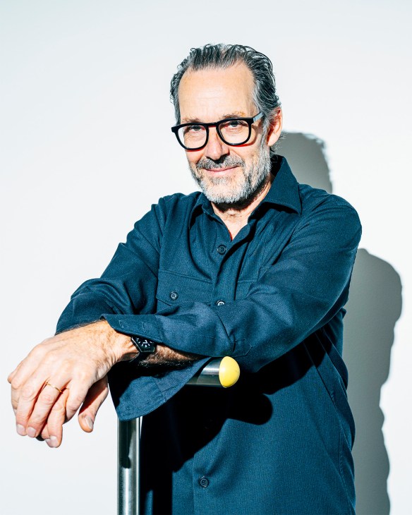 Portrait of Konstantin Grcic, a bespectacled man with gray hair and beard wearing a dark blue shirt, leaning on a cane with a yellow knob.