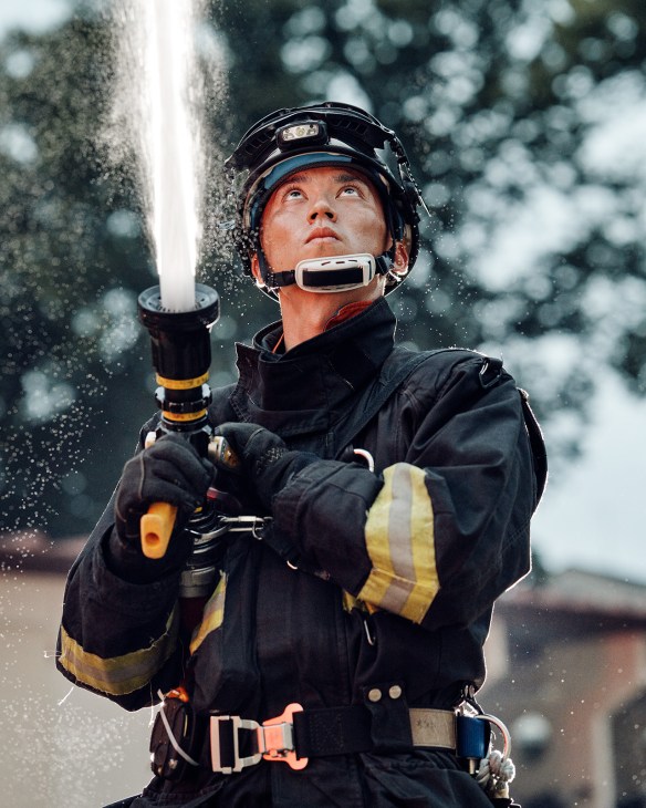 Tokyo Fire Department firefighter in black hazmat suit with yellow reflective stripes holding pressurized water hose while training.