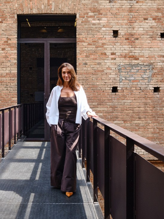 Woman in white blazer and black pants standing on brick building entryway with metal railings.