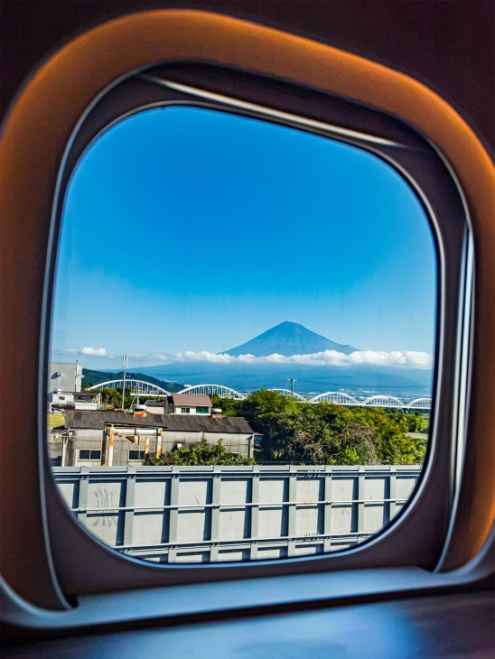 View of snow-capped Mount Fuji framed by an airplane window with tea gardens and buildings visible below.
