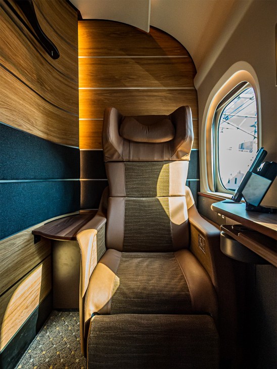 Interior view of a Tokaido Shinkansen Supreme Class private cabin with brown leather seat, wooden paneling, and window showing airport tarmac outside.