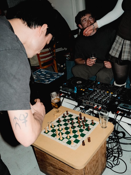 People gather around a chessboard at Acid Chess Club in Amsterdam, with a DJ mixing music and drinks in hand.