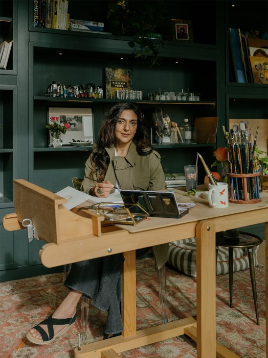 Creative director Tara Lal sits at a wooden desk in her book-lined studio with paintbrushes and art supplies.