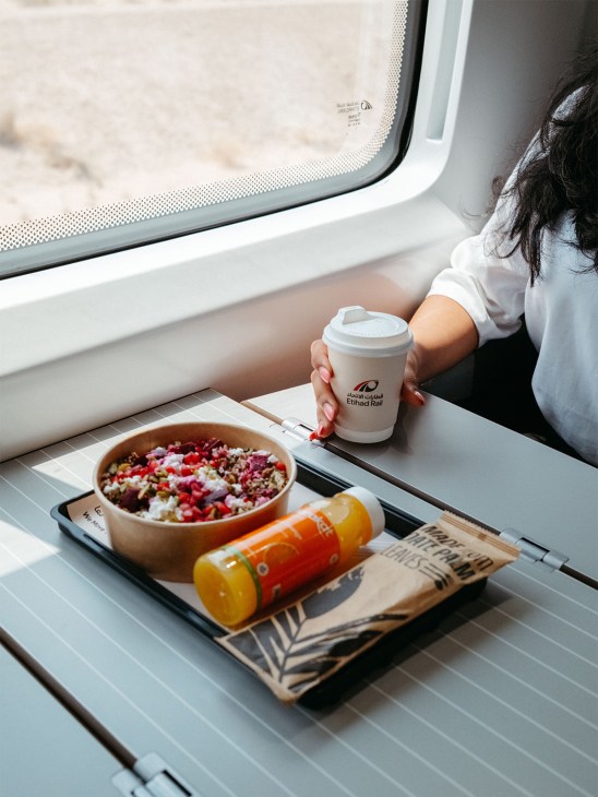 Passenger holding Etihad Rail coffee cup by train window with tray of colorful food and beverages.