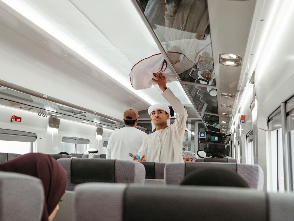 A train attendant in white uniform retrieves luggage from the overhead compartment on an Etihad Rail passenger train.